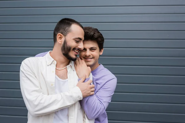 Усміхнений брюнетка гей чоловік в сорочці і брекети обіймає партнера і дивиться на камеру, стоячи разом біля сірого будинку на вулиці міста — стокове фото