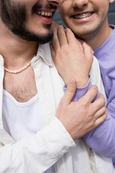 Cropped view of carefree homosexual man in purple sweatshirt with braces hugging and touching shoulder of bearded partner outdoors at daytime — Stock Photo