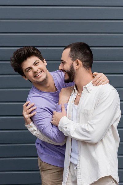 Portrait of cheerful homosexual man in purple sweatshirt and braces hugging young bearded boyfriend and looking at camera while standing near building outdoors — Stock Photo