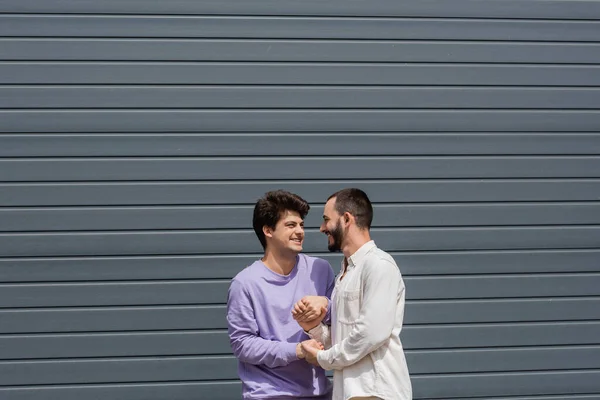Positive and young homosexual couple holding hands while talking and looking at each other near building on urban street at daytime — Stock Photo