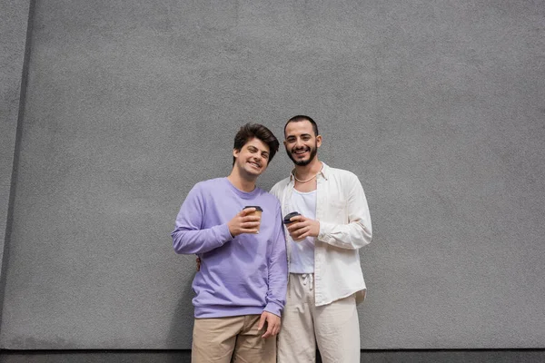 Улыбающиеся и молодые однополые пары в повседневной одежде, держащие кофе, чтобы пойти и посмотреть в камеру, стоя рядом со зданием — стоковое фото