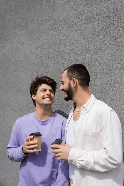 Bearded homosexual man holding coffee to go and looking at smiling boyfriend in braces while talking near building on urban street at daytime — Stock Photo