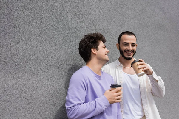 Glücklicher bärtiger homosexueller Mann mit Kaffee in der Hand in lila Sweatshirt und Hosenträgern, während er in der Nähe eines Gebäudes auf der städtischen Straße steht — Stockfoto