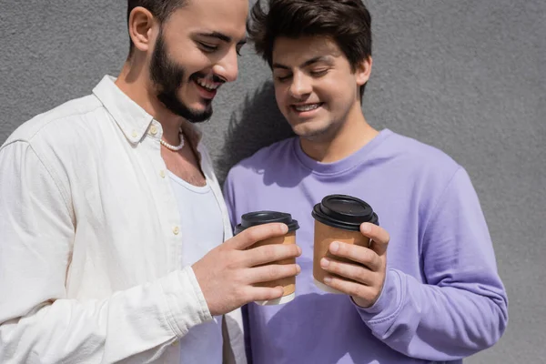 Young carefree same sex couple in casual clothes holding takeaway coffee in paper cups while standing near building outdoors at daytime — Stock Photo