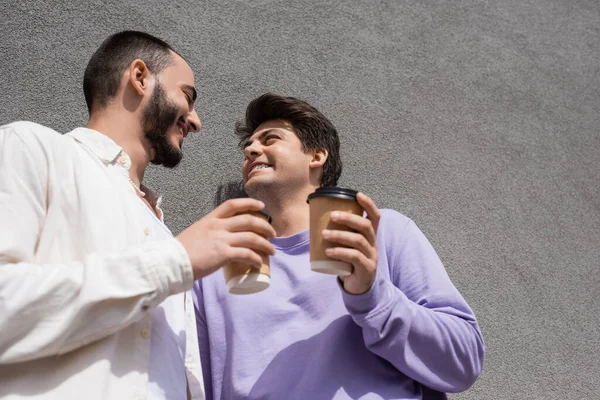 Низкий угол зрения улыбки и молодые однополые пары в повседневной одежде держа кофе, чтобы пойти, стоя рядом со зданием на открытом воздухе — стоковое фото