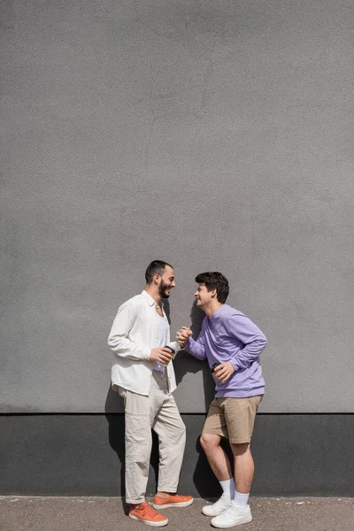 Вид сбоку улыбающихся однополых партнеров, держащихся за руки и кофе, чтобы пойти в бумажных чашках, стоя у стены на городской улице в дневное время — стоковое фото