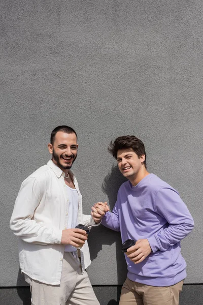 Positives und junges gleichgeschlechtliches Paar, das Händchen hält und Kaffee zum Mitnehmen trinkt, während es tagsüber in die Kamera in der Nähe eines grauen Gebäudes blickt — Stockfoto