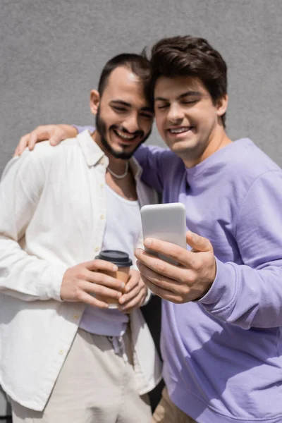 Размытый гей обнимает улыбающегося парня в повседневной одежде с кофе, чтобы пойти и использовать мобильный телефон рядом с серым зданием на открытом воздухе — стоковое фото