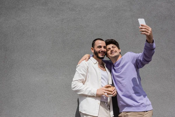 Молодая и веселая однополые пара с кофе, чтобы пойти обниматься и делать селфи на смартфоне, стоя возле здания в дневное время — стоковое фото