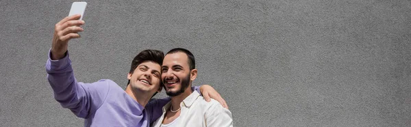 Allegro coppia omosessuale in abiti casual prendendo selfie su smartphone insieme mentre in piedi vicino al muro grigio edificio sulla strada urbana di giorno, banner — Foto stock