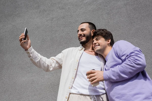 Vista de ángulo bajo de hombre gay positivo y barbudo tomando selfie con novio despreocupado sosteniendo café para llevar cerca del edificio al aire libre durante el día - foto de stock