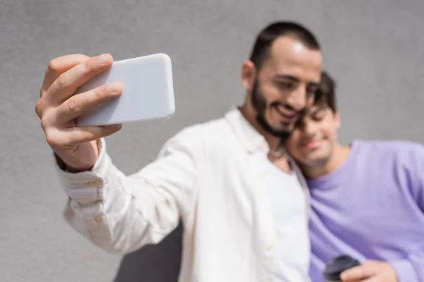 Blurred same sex couple in casual clothes taking selfie on smartphone together while standing near building wall on urban street at daytime — Stock Photo