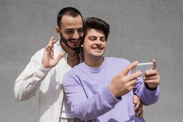 Улыбающаяся гей-пара в повседневной одежде с видеозвонком на размытый смартфон, стоя рядом со зданием на городской улице днем — стоковое фото