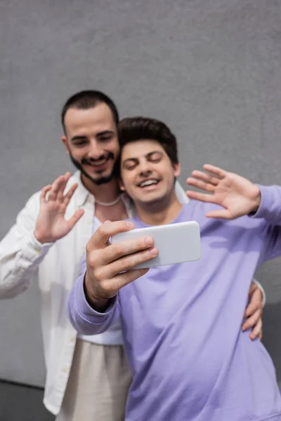 Blurred and young same sex boyfriends in casual clothes having video call on cellphone while standing together near building outdoors — Stock Photo