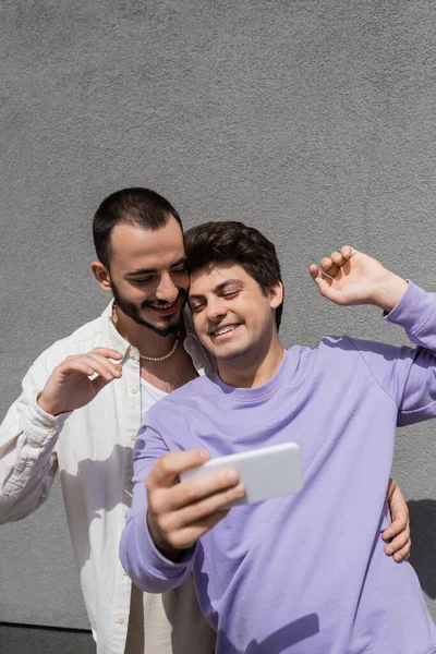 Веселый гомосексуалист в повседневной одежде с помощью мобильного телефона, стоя рядом с парнем в брекетах и здании на городской улице — стоковое фото