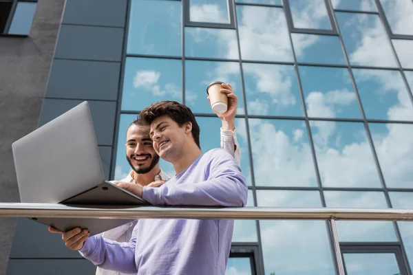 Blick auf ein lächelndes und junges homosexuelles Paar, das einen Coffee to go in der Hand hält und einen Laptop benutzt, während es in der Nähe eines verschwommenen Gebäudes auf der städtischen Straße steht — Stockfoto