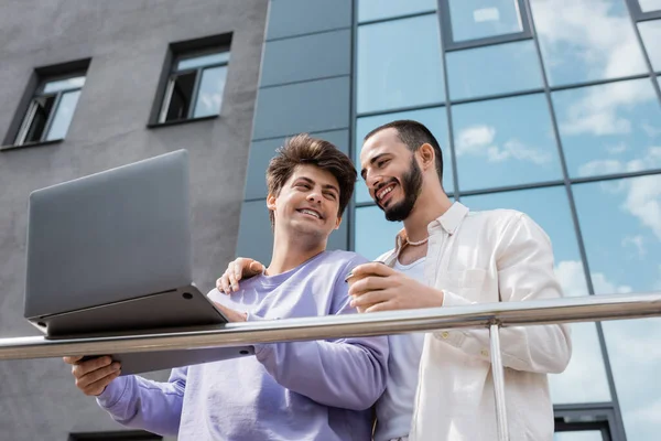 Blick auf ein fröhliches homosexuelles Paar in lässiger Kleidung mit Coffee to go und Laptop in der Nähe von Geländern und unscharfen Gebäuden im Freien, auf der städtischen Straße — Stockfoto