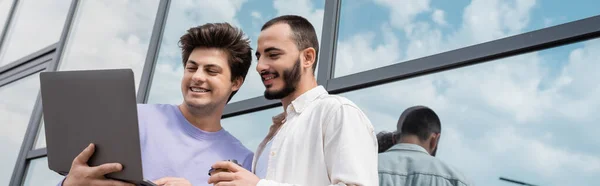 Young and cheerful same sex couple with takeaway coffee in paper cup using laptop while standing together near building on urban street, banner — Stock Photo