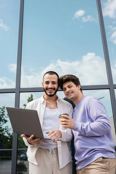 Jovem e alegre casal homossexual com café para ir e laptop sorrindo para a câmera enquanto está perto de edifício na rua urbana, fora — Fotografia de Stock