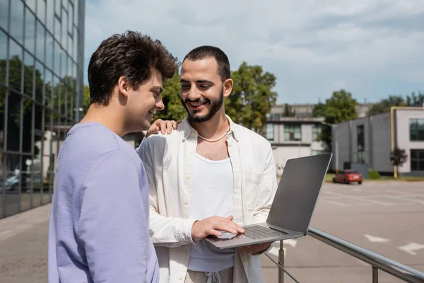 Positive gay man in casual clothes hugging bearded boyfriend using laptop with blank screen near blurred building on urban street at daytime — Stock Photo