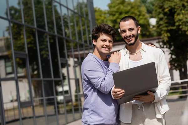 Молода і безтурботна гомосексуальна пара в повсякденному одязі тримає ноутбук і посміхається на камеру, стоячи біля розмитої будівлі на міській вулиці — стокове фото