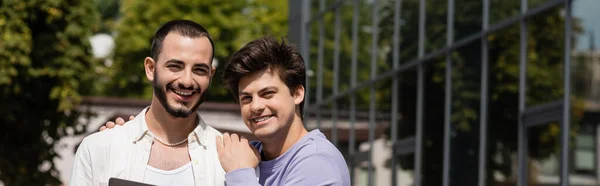 Улыбающийся гомосексуалист в повседневной одежде обнимает бородатого бойфренда и смотрит вместе в камеру на улице днем, на городской улице, баннер — стоковое фото