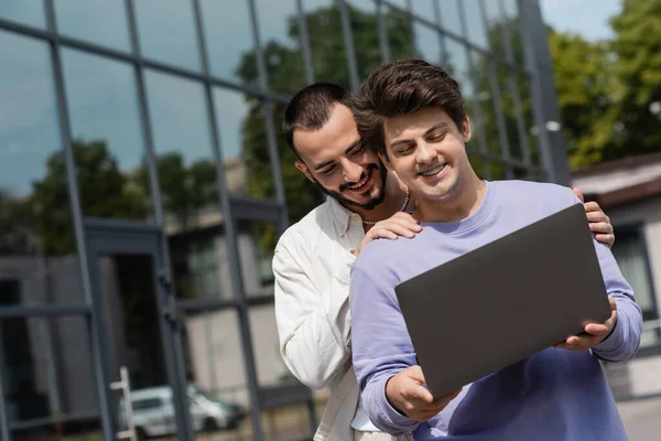 Overjoyed and bearded man hugging smiling boyfriend with braces in casual clothes using laptop while standing together near blurred building on urban street — Stock Photo