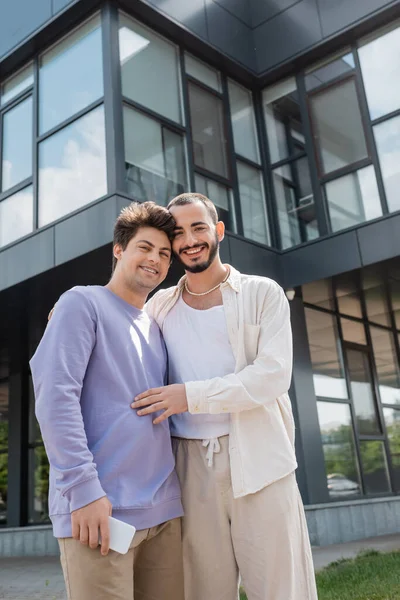 Couple homosexuel souriant et jeune en vêtements décontractés avec smartphone regardant la caméra tout en s'embrassant dans la rue urbaine le jour — Photo de stock