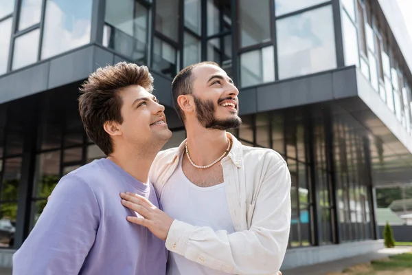 Hombre homosexual sonriente abrazando al joven novio con sudadera y tirantes mientras miran hacia otro lado juntos cerca de un edificio borroso en la calle urbana durante el día - foto de stock