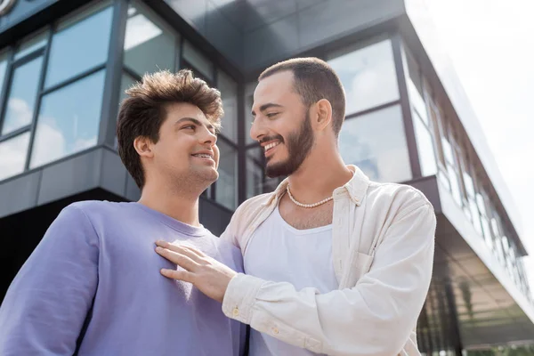 Vista de ángulo bajo de las parejas del mismo sexo despreocupadas en ropa casual mirándose mientras están de pie cerca de un edificio borroso en la calle urbana durante el día — Stock Photo