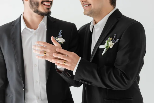 Vista recortada del novio gay sonriente en elegantes trajes con boutonniere floral tocando la mano del novio en tirantes con anillo de bodas mientras está de pie aislado en gris - foto de stock
