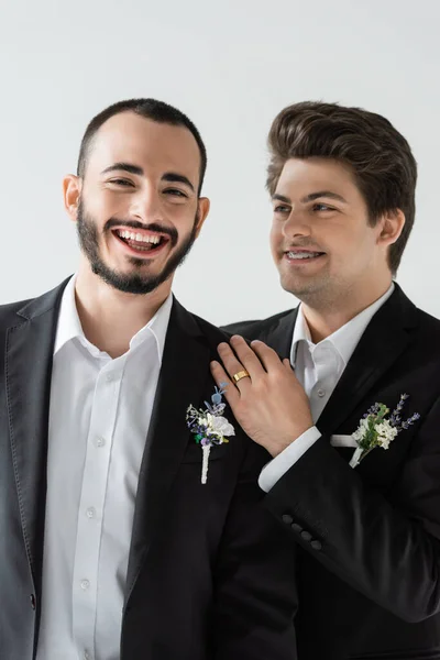 Smiling brunette gay man in elegant suit and braces hugging and looking at bearded boyfriend with boutonniere during wedding celebration isolated on grey — Stock Photo