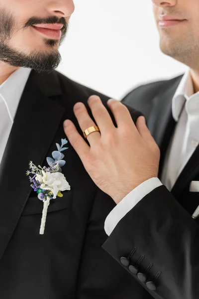 Cropped view of blurred gay groom in elegant suit with floral boutonniere and wedding ring on hand hugging bearded boyfriend while standing isolated on grey — Stock Photo