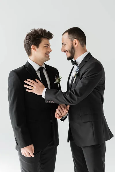 Alegre e barbudo gay homem no elegante formal desgaste com floral boutonniere segurando mão de noivo durante casamento celebração isolado no cinza — Fotografia de Stock