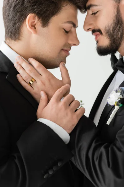 Vue latérale de jeunes mariés de même sexe en costumes classiques avec des boutonnières se touchant lors de la célébration du mariage et tout en se tenant isolé sur gris — Photo de stock