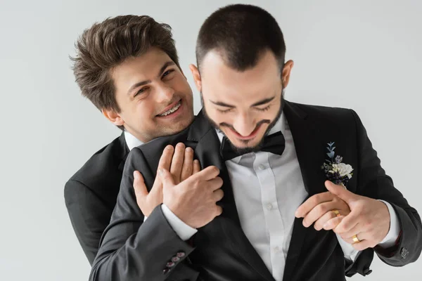 Portrait of positive homosexual man with braces in elegant suit and golden ring on hand hugging bearded groom during wedding celebration isolated on grey — Stock Photo