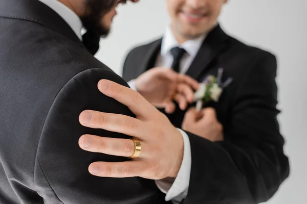Обрізаний вид розмитого гей-чоловіка із золотим кільцем на руці, який торкається плеча партнера в класичному костюмі під час весільного торжества ізольовано на сірому — стокове фото