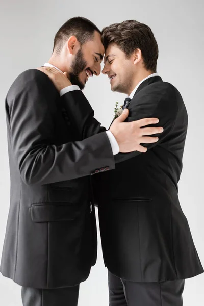 Vista lateral de la sonriente pareja del mismo sexo con los ojos cerrados en trajes que se tocan durante la celebración de la boda mientras están de pie aislados en gris - foto de stock