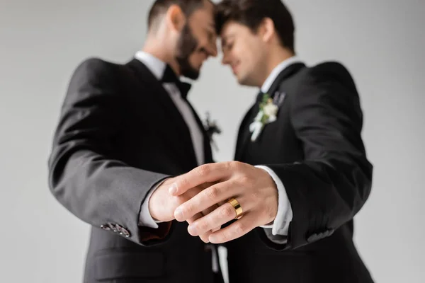 Side view of blurred same sex couple in formal wear with boutonnieres touching hands of each other with golden ring during wedding isolated on grey — Stock Photo