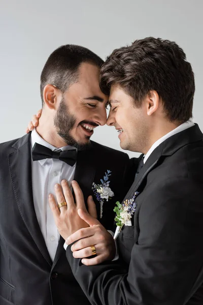 Portrait de jeune homme gay avec bretelles en costume élégant avec boutonnière touchant petit ami positif et barbu lors de la cérémonie de mariage isolé sur gris — Photo de stock