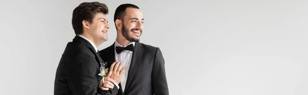 Позитивний гомосексуальний чоловік у костюмі з бутоньєрою, який торкається бородатого хлопця і дивиться в сторону під час весільної церемонії ізольовано на сірому, банер — стокове фото