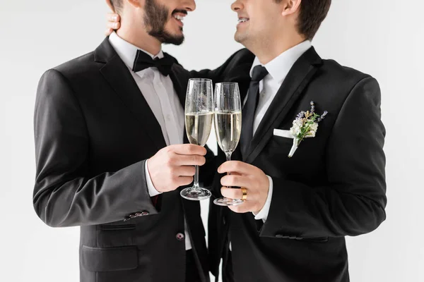 Обрезанный вид улыбающегося гомосексуального жениха в брекетах и костюме с бутоньеркой обнимающей бородатого бойфренда и тостом с шампанским во время свадебной церемонии, изолированной на серой — стоковое фото