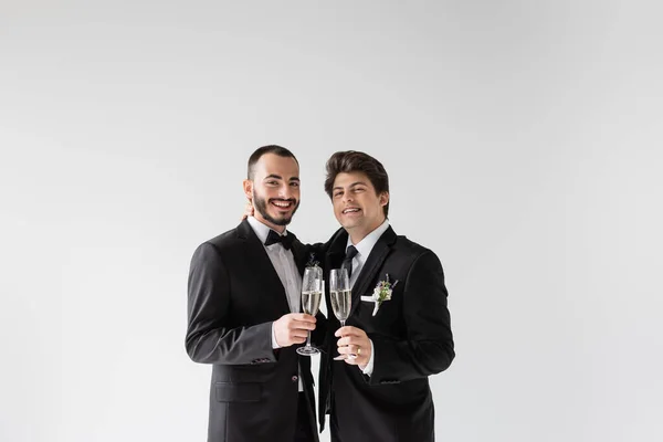 Улыбающаяся и молодая однополая пара в элегантных костюмах с цветочными бутоньерными объятиями и бокалами шампанского во время свадебной церемонии, изолированной на сером — стоковое фото