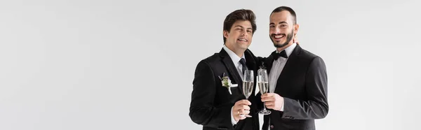 Positives gleichgeschlechtliches Paar in eleganten Anzügen mit Champagnergläsern und Blick in die Kamera während der Hochzeitszeremonie isoliert auf grauem Banner — Stockfoto