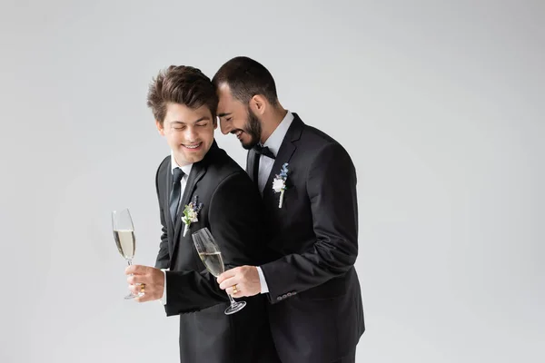 Улыбающийся и бородатый гей-жених в элегантной формальной одежде с бутоньеркой, держа бокал шампанского рядом с бойфрендом в брекетах с закрытыми глазами во время свадебного торжества, изолированного на сером — стоковое фото