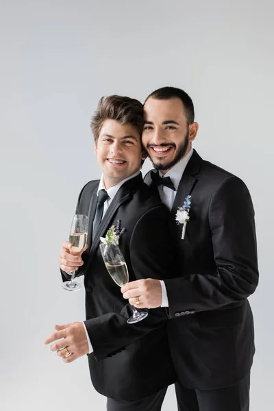Portrait of positive same sex couple in elegant classic suits holding glasses of champagne while standing together during wedding celebration isolated on grey — Stock Photo