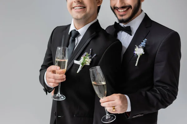 Cropped view of smiling homosexual grooms in classic attire with floral boutonnieres holding glasses of champagne while celebrating wedding isolated on grey — Stock Photo
