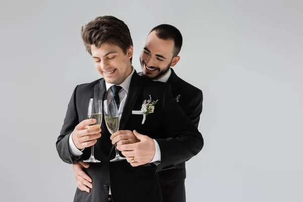 Улыбающийся и бородатый гомосексуальный жених трогает руку элегантного бойфренда в костюме, держа бокалы шампанского во время свадебного торжества, изолированного на сером — стоковое фото