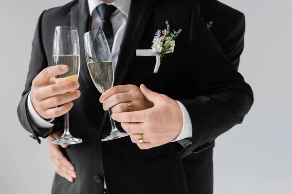Обрезанный вид гей-пары в классических костюмах и золотых кольцах с бокалами шампанского во время празднования свадьбы изолированы на сером — стоковое фото