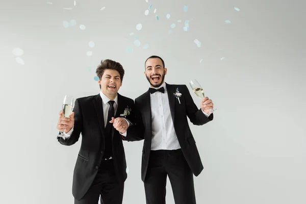 Excited same sex grooms in classic suits with boutonnieres holding hands and glasses of champagne while standing under falling confetti during wedding celebration on grey background — Stock Photo